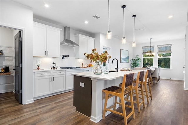 kitchen featuring pendant lighting, white cabinetry, a kitchen bar, a kitchen island with sink, and wall chimney range hood