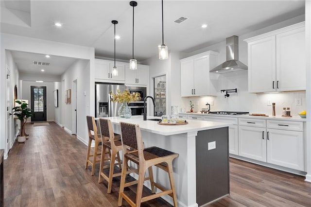 kitchen featuring stainless steel fridge with ice dispenser, white cabinets, decorative light fixtures, a center island with sink, and wall chimney exhaust hood