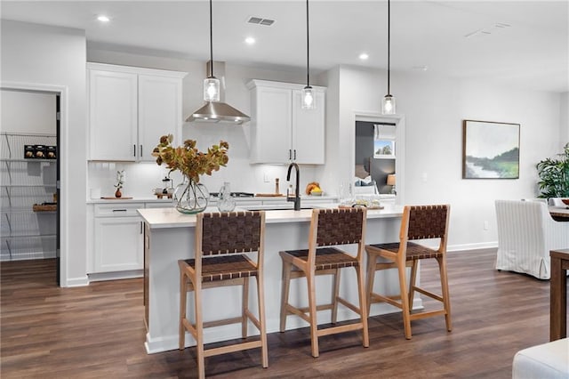 kitchen with decorative light fixtures, white cabinetry, wall chimney range hood, dark hardwood / wood-style floors, and a kitchen island with sink