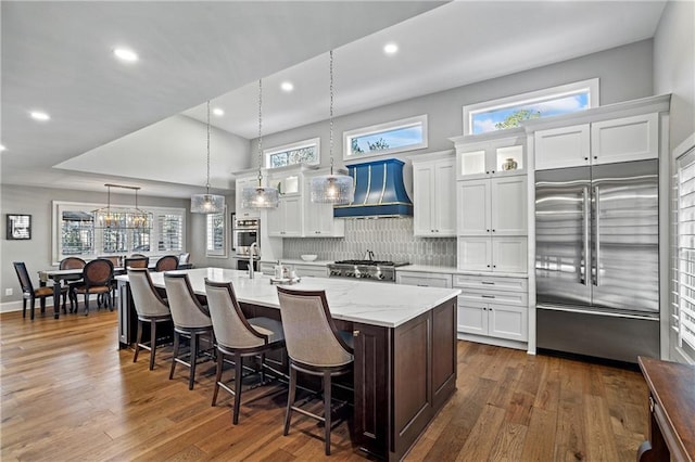 kitchen featuring premium range hood, a kitchen island with sink, hanging light fixtures, white cabinetry, and stainless steel appliances