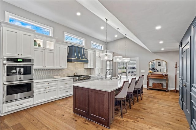 kitchen with premium range hood, lofted ceiling, white cabinetry, a center island with sink, and stainless steel appliances