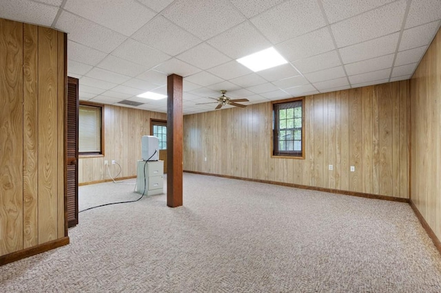 basement with carpet flooring, a ceiling fan, baseboards, a drop ceiling, and wooden walls
