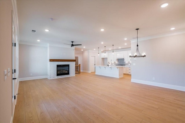 unfurnished living room with light wood-type flooring, ornamental molding, a brick fireplace, sink, and ceiling fan with notable chandelier