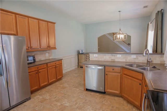 kitchen featuring a wainscoted wall, stainless steel appliances, dark countertops, hanging light fixtures, and a sink