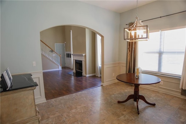 unfurnished dining area with wainscoting, a fireplace with flush hearth, stairs, a chandelier, and a decorative wall