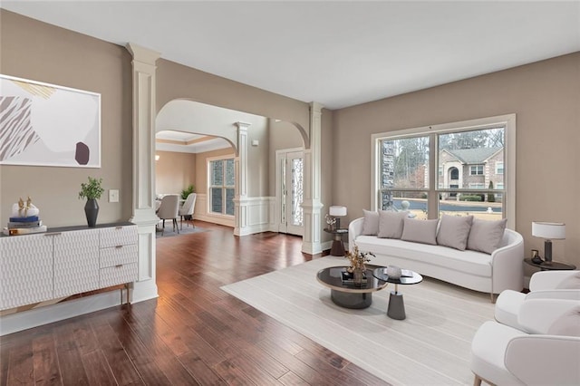 living area featuring arched walkways, a decorative wall, wood finished floors, wainscoting, and decorative columns