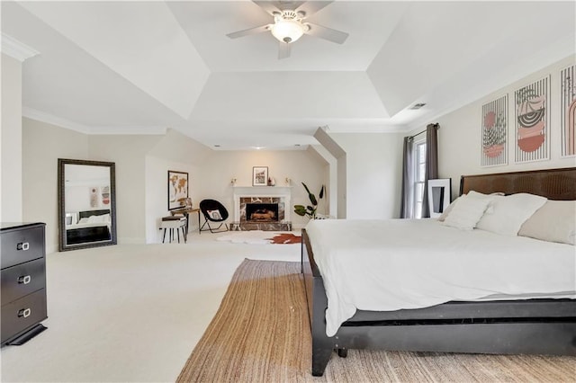 carpeted bedroom featuring a ceiling fan, visible vents, a tray ceiling, a fireplace with raised hearth, and crown molding