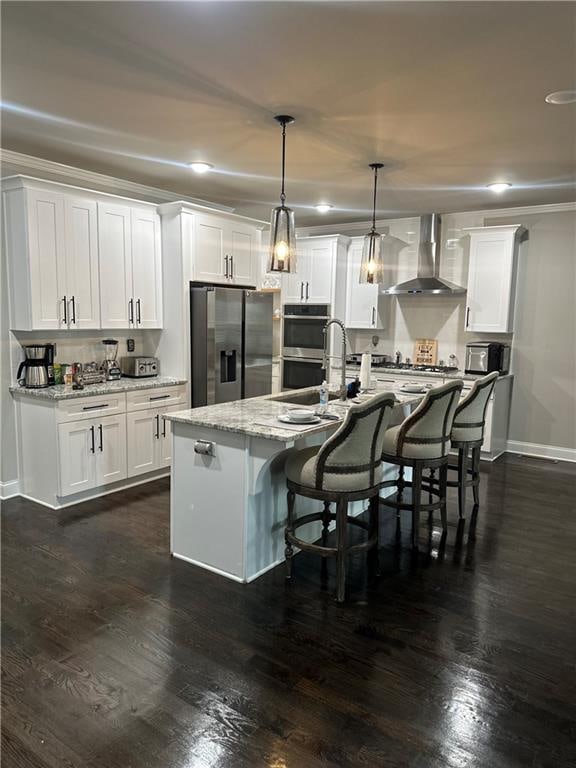 kitchen with white cabinetry, wall chimney exhaust hood, hanging light fixtures, a center island with sink, and appliances with stainless steel finishes