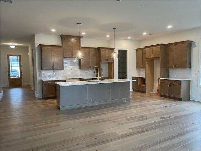 kitchen featuring sink, light hardwood / wood-style flooring, pendant lighting, a kitchen island with sink, and backsplash