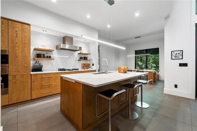 kitchen with an island with sink, decorative light fixtures, sink, and wall chimney range hood