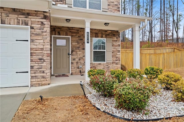 doorway to property featuring a garage, stone siding, covered porch, and fence