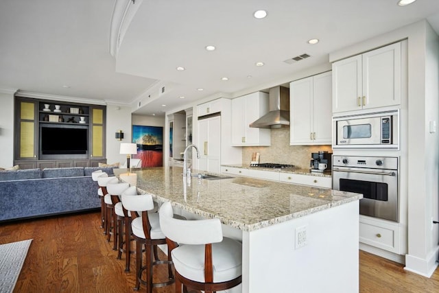 kitchen with wall chimney range hood, a sink, built in appliances, ornamental molding, and visible vents