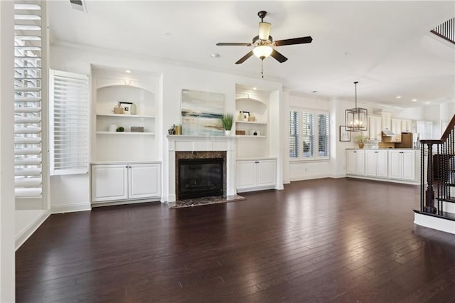 unfurnished living room featuring stairs, dark wood finished floors, a fireplace, ceiling fan with notable chandelier, and built in shelves