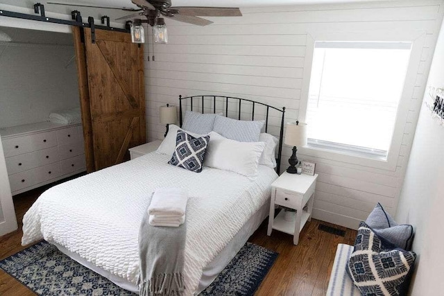 bedroom with a barn door, ceiling fan, dark wood-type flooring, and wood walls