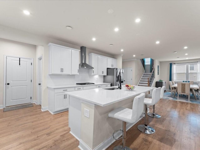 kitchen with a sink, light wood-style floors, wall chimney range hood, an island with sink, and stainless steel fridge