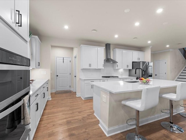 kitchen with light wood-style flooring, a sink, appliances with stainless steel finishes, wall chimney exhaust hood, and a center island with sink