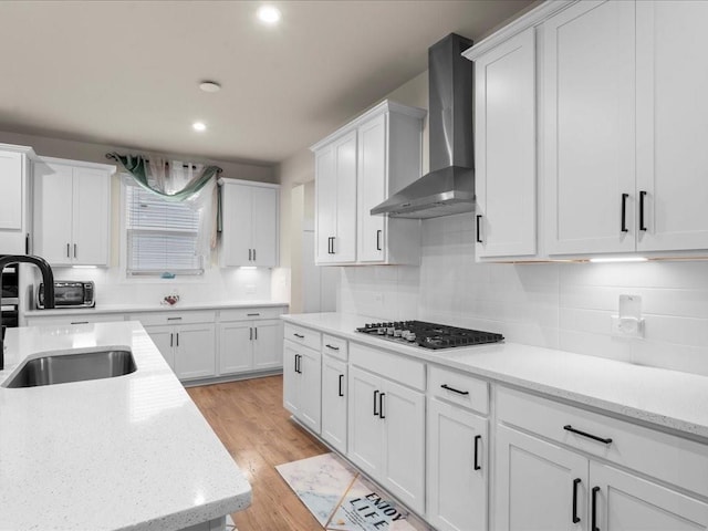 kitchen with stainless steel gas cooktop, light wood finished floors, white cabinetry, a sink, and wall chimney range hood