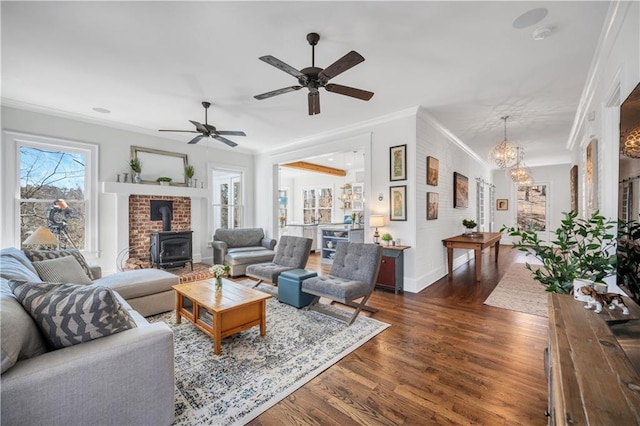 living area with a wood stove, dark wood finished floors, ceiling fan with notable chandelier, baseboards, and ornamental molding