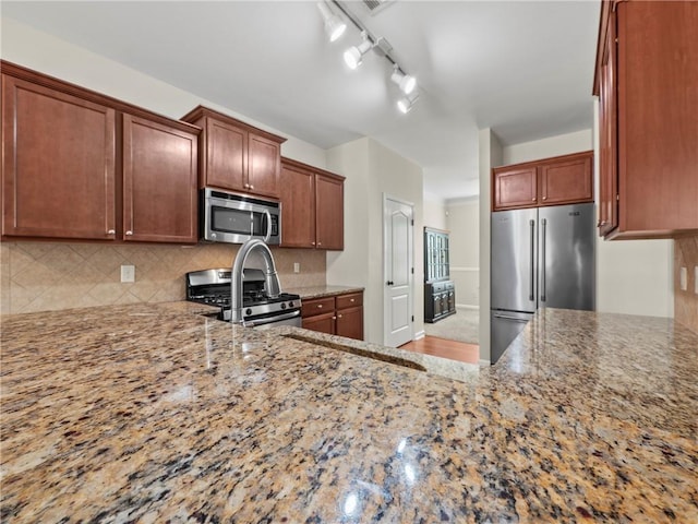 kitchen with light stone counters, brown cabinets, backsplash, appliances with stainless steel finishes, and a peninsula