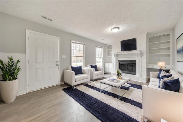 living room with a brick fireplace, built in shelves, wood-type flooring, and a textured ceiling