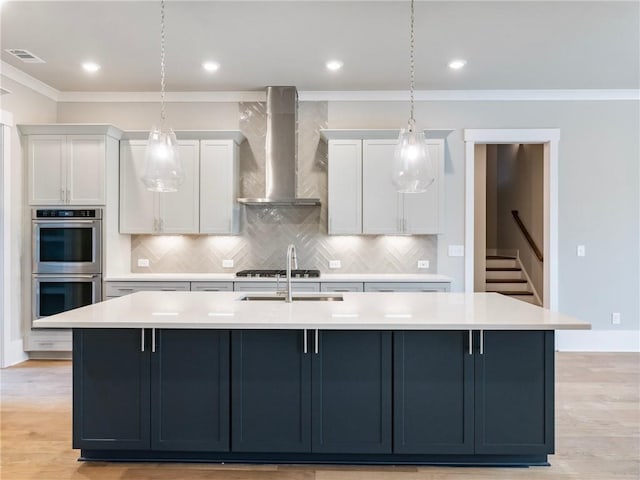 kitchen with appliances with stainless steel finishes, wall chimney range hood, decorative light fixtures, white cabinets, and an island with sink