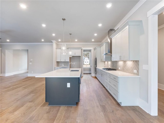 kitchen with wall chimney exhaust hood, sink, decorative light fixtures, light hardwood / wood-style floors, and an island with sink