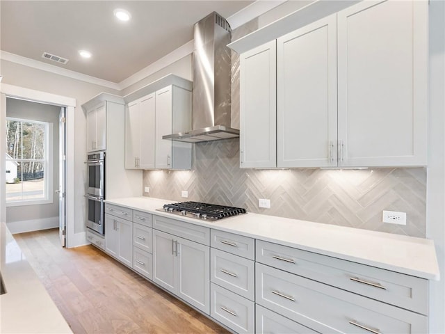 kitchen with wall chimney exhaust hood, light hardwood / wood-style flooring, backsplash, appliances with stainless steel finishes, and ornamental molding