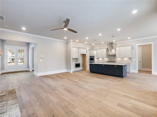 kitchen featuring wall chimney range hood, light hardwood / wood-style floors, decorative light fixtures, a center island with sink, and white cabinets