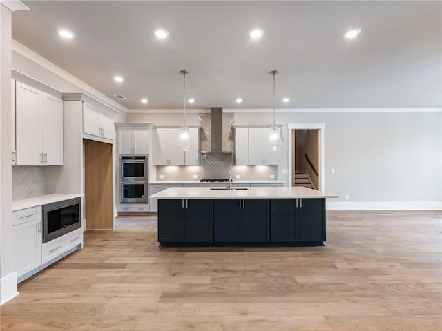 kitchen featuring wall chimney range hood, an island with sink, pendant lighting, white cabinets, and appliances with stainless steel finishes