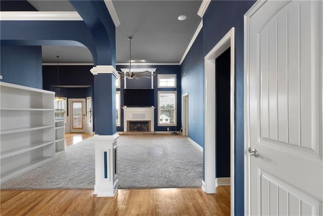 unfurnished living room featuring crown molding, a fireplace with raised hearth, a ceiling fan, wood finished floors, and ornate columns