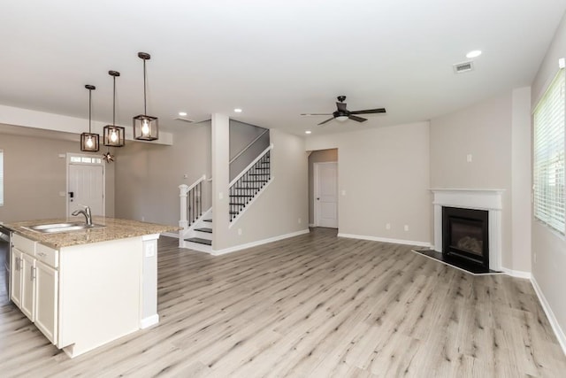 kitchen featuring light wood-type flooring, light stone counters, a kitchen island with sink, decorative light fixtures, and ceiling fan