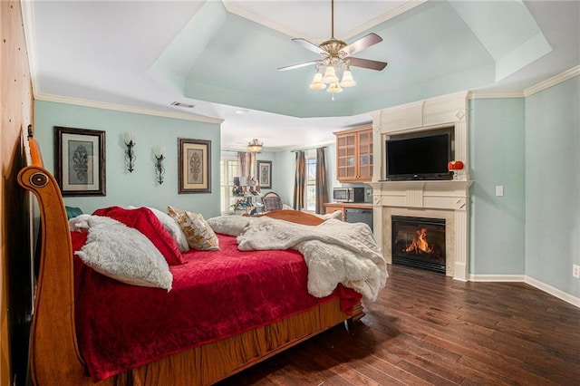 bedroom featuring baseboards, a raised ceiling, ornamental molding, wood-type flooring, and a glass covered fireplace