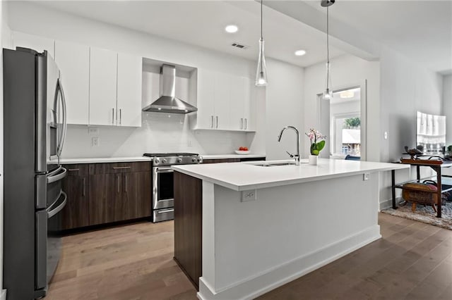 kitchen featuring sink, an island with sink, dark brown cabinetry, wall chimney range hood, and appliances with stainless steel finishes