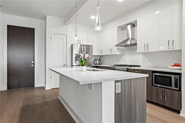 kitchen with white cabinetry, hanging light fixtures, wall chimney range hood, a kitchen island with sink, and appliances with stainless steel finishes