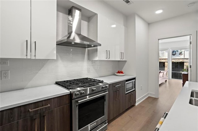 kitchen featuring white cabinetry, hardwood / wood-style floors, dark brown cabinetry, wall chimney range hood, and appliances with stainless steel finishes