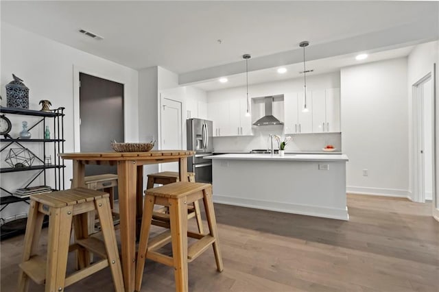 kitchen featuring white cabinetry, light wood-type flooring, stainless steel refrigerator with ice dispenser, wall chimney range hood, and pendant lighting