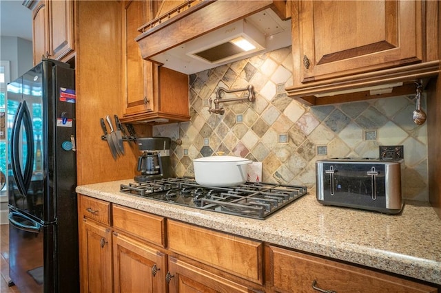 kitchen featuring light stone counters, stainless steel gas stovetop, decorative backsplash, black refrigerator, and custom range hood