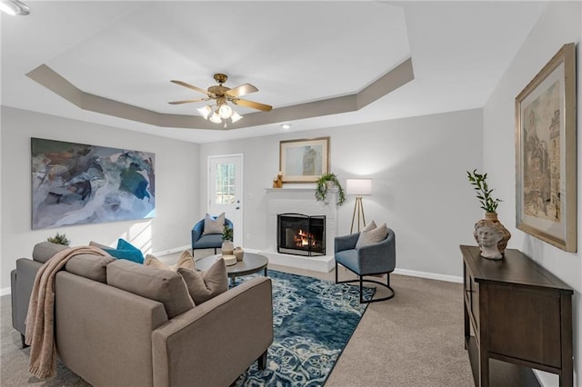 living area featuring a brick fireplace, baseboards, carpet floors, a raised ceiling, and a ceiling fan