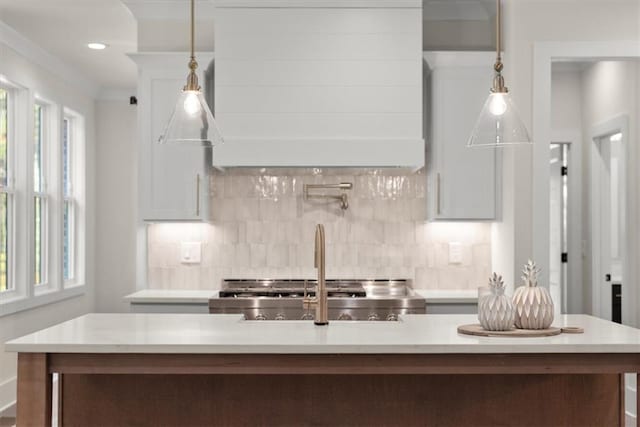 kitchen with decorative backsplash, custom range hood, white cabinets, hanging light fixtures, and an island with sink