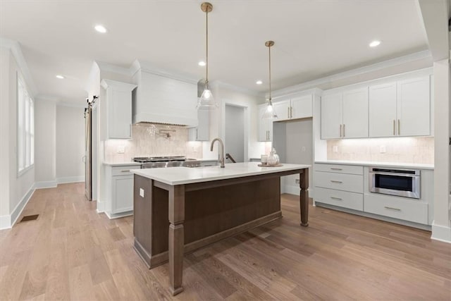 kitchen with hanging light fixtures, crown molding, an island with sink, light hardwood / wood-style floors, and white cabinets