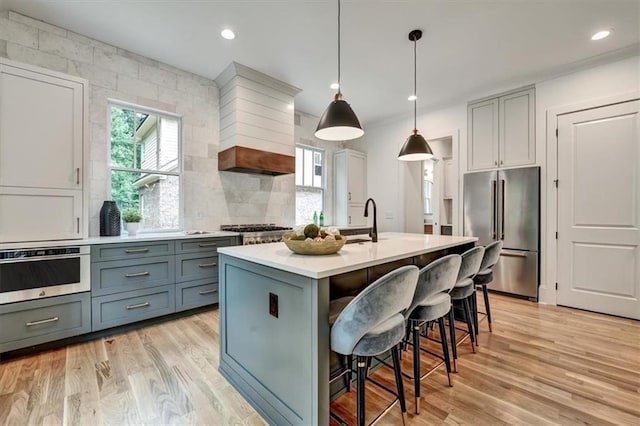 kitchen with custom range hood, a kitchen island with sink, appliances with stainless steel finishes, and light hardwood / wood-style flooring