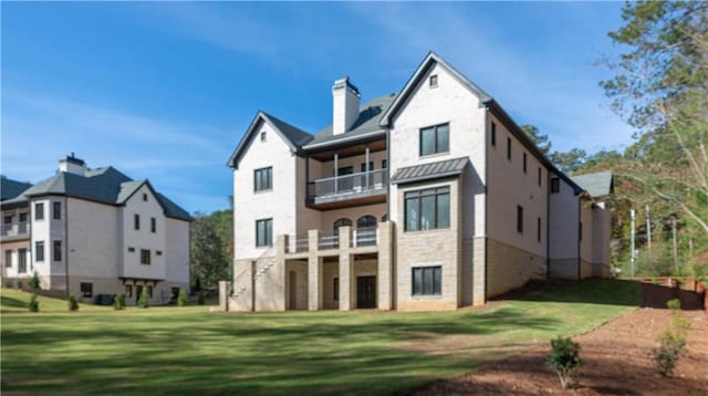 back of house featuring metal roof, a balcony, a lawn, a standing seam roof, and a chimney