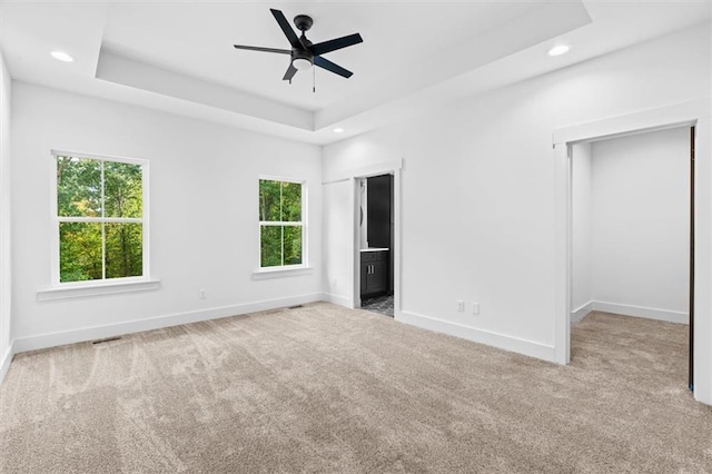 unfurnished bedroom with a raised ceiling, multiple windows, light colored carpet, and ceiling fan