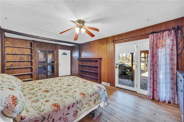 bedroom featuring hardwood / wood-style flooring, wood walls, ceiling fan, access to exterior, and a textured ceiling