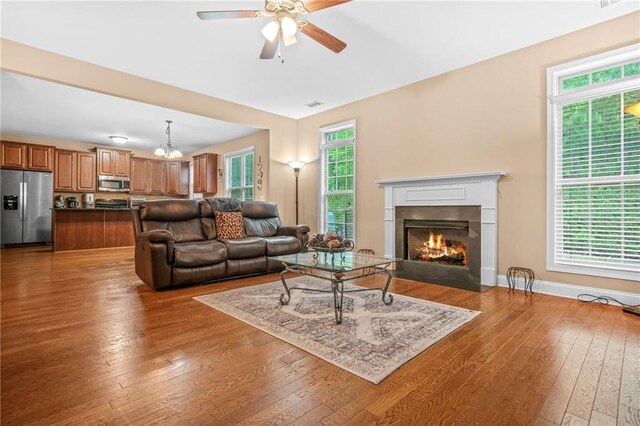 living room featuring wood-type flooring, visible vents, a fireplace with flush hearth, baseboards, and a ceiling fan