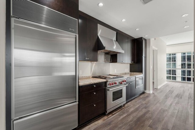 kitchen with premium appliances, light stone countertops, wall chimney exhaust hood, wood-type flooring, and dark brown cabinetry