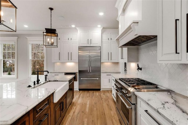 kitchen featuring sink, high end appliances, white cabinetry, light stone counters, and hanging light fixtures