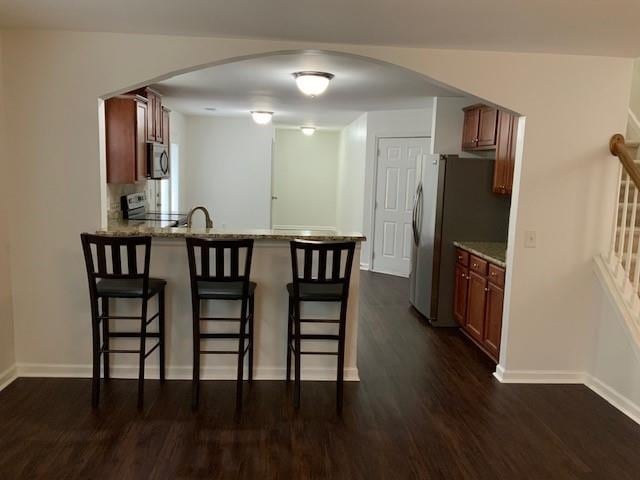 kitchen with stainless steel appliances, kitchen peninsula, light stone counters, a kitchen bar, and dark wood-type flooring