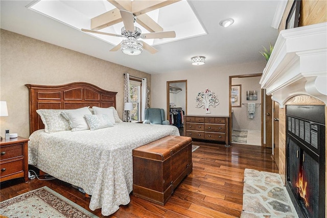 bedroom with a walk in closet, a lit fireplace, wood-type flooring, and a ceiling fan