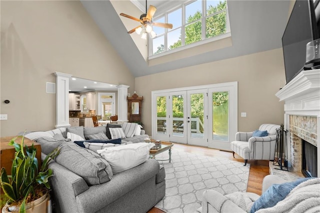 living area featuring light wood-style flooring, decorative columns, a ceiling fan, a brick fireplace, and high vaulted ceiling